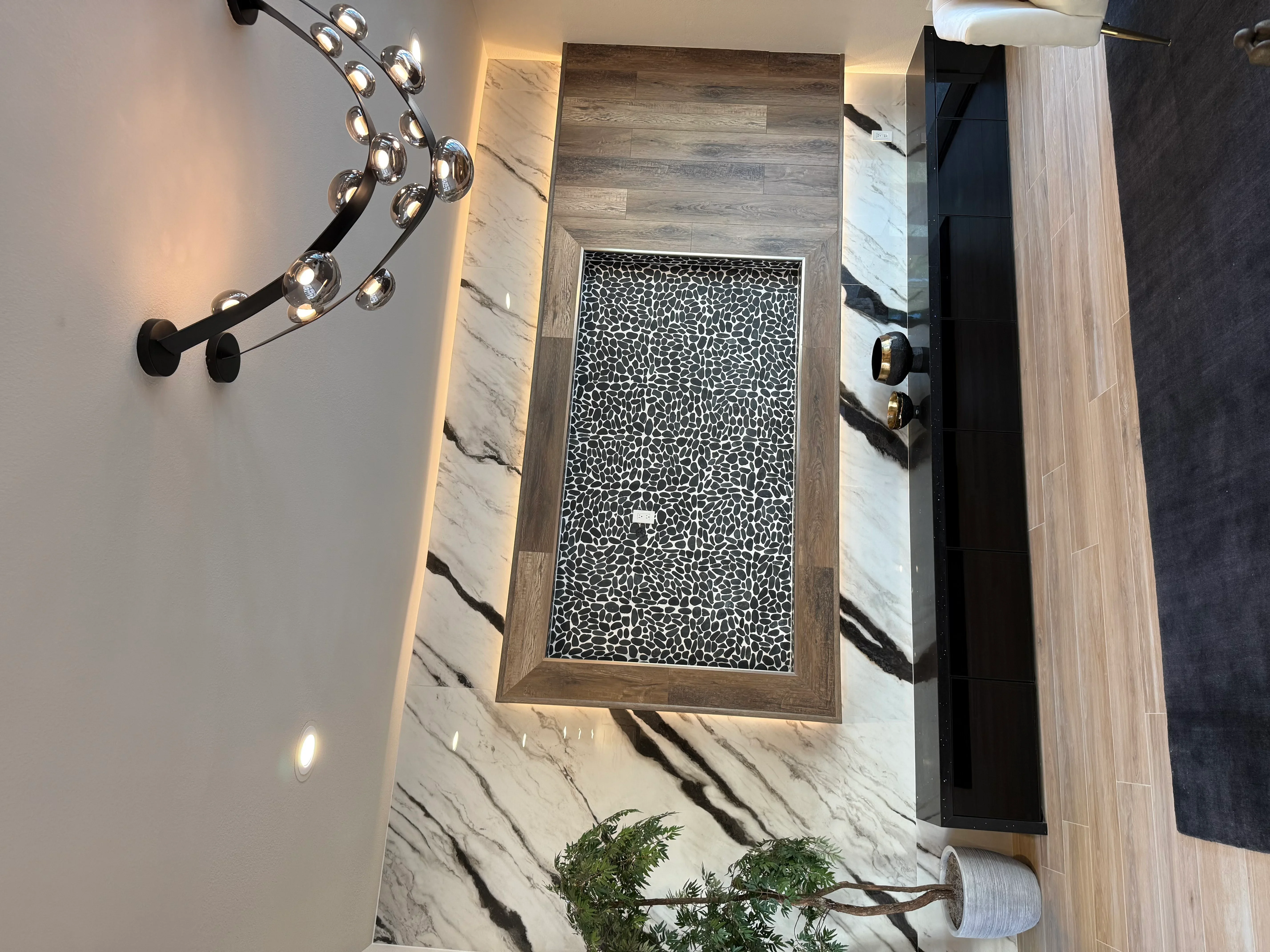 Marble tile bathroom wall with black pebble mosaic inset, wood-frame surround, and chrome pendant lighting