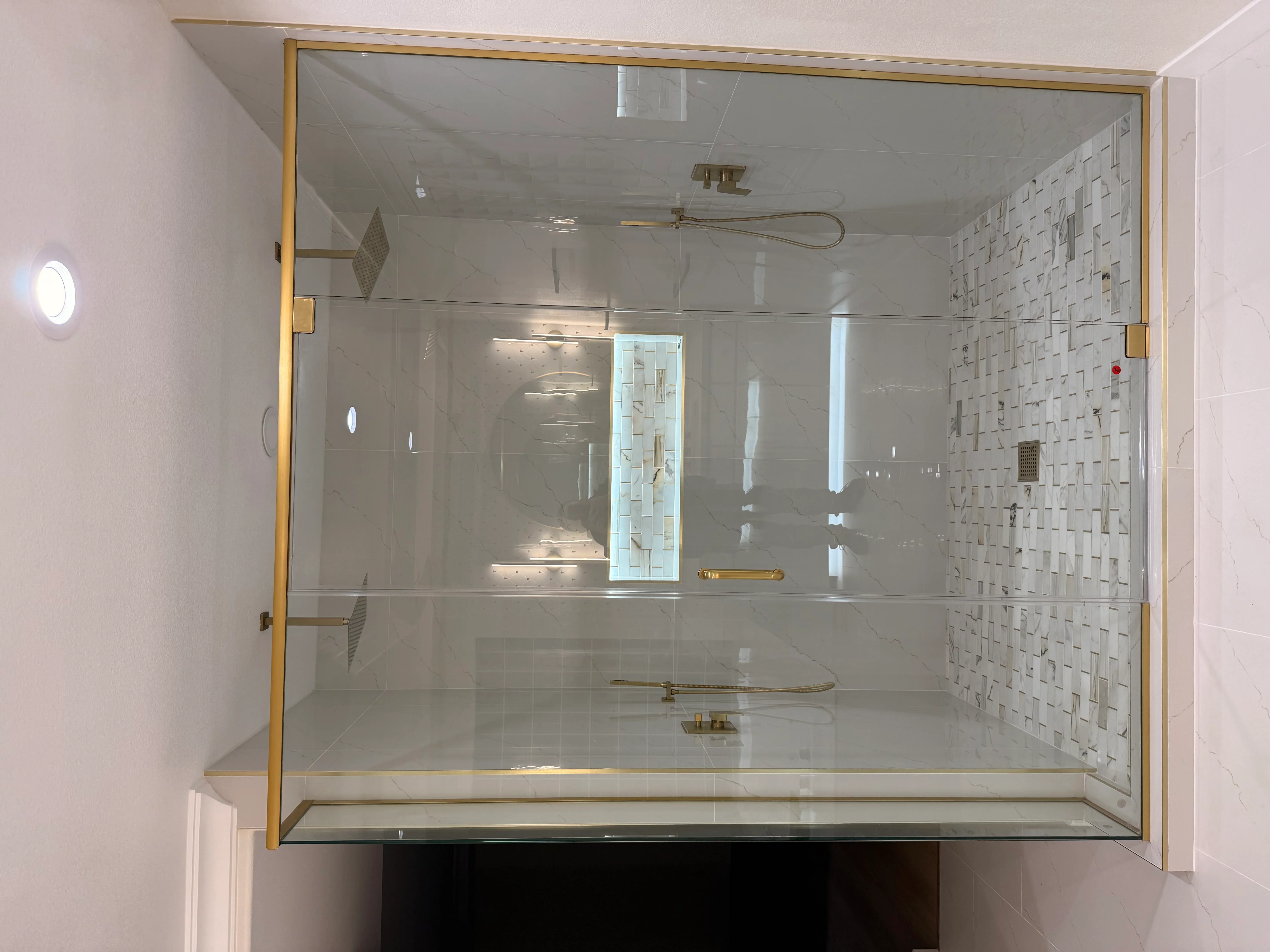 Frameless glass shower enclosure with brushed gold hardware, textured tile walls, and rainfall showerhead