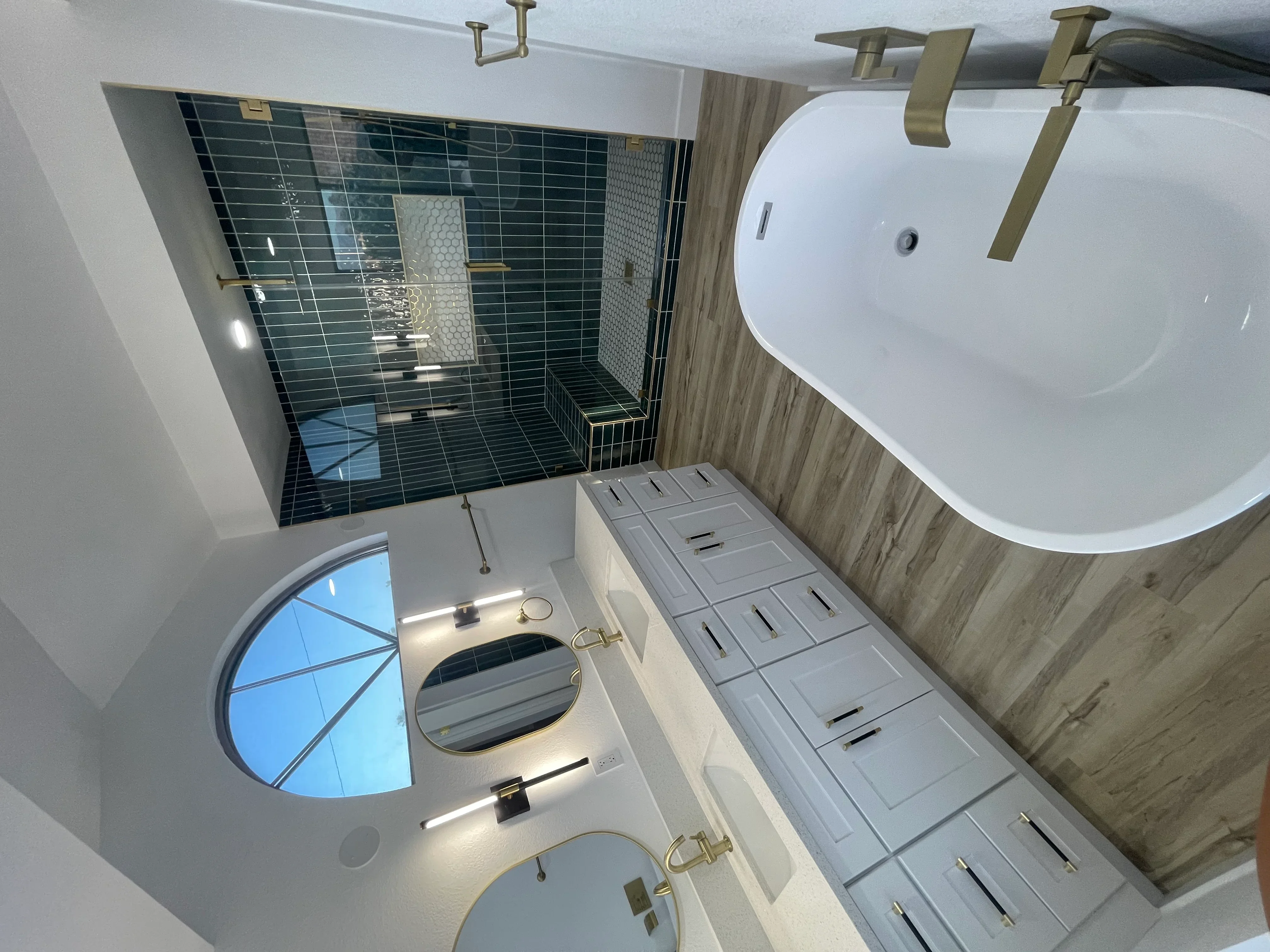 Bathroom with dark green tile shower, freestanding tub, dual vanities, brass fixtures, and wood-look walls